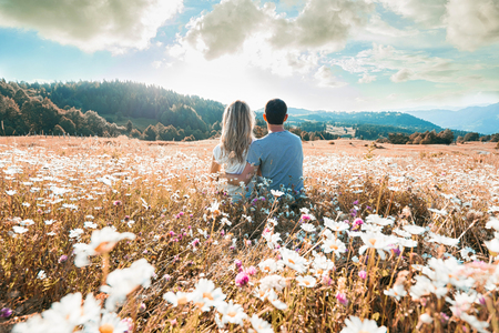 Young couple sitting on the chamomiles field and looking at the cloudy sky. Back view. Фото со стока