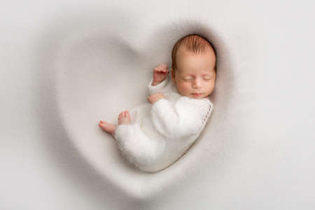 Un garçon nouveau-né endormi dans les premiers jours de la vie dans un cocon doux blanc avec un bonnet blanc en laine tricoté sur fond blanc en forme de coeur. macrophotographie en studio, portrait d'un nouveau-né. Banque d'images