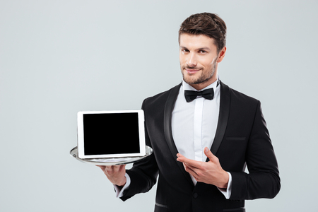 Confident young butler in tuxedo holding and pointing at blank screen tablet on tray Фото со стока