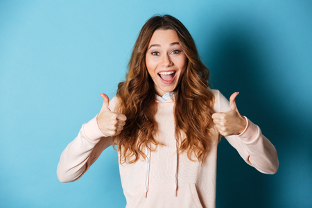 Image of cute happy lady standing isolated over blue wall background. Looking camera showing thumbs up..