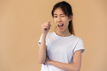 Photo of cute korean woman wearing basic t-shirt smiling and winking at you isolated over beige background in studio