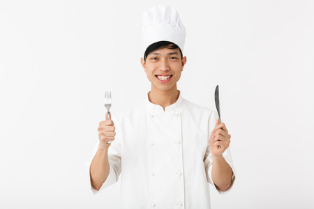 Image of asian young chief man in white cook uniform smiling at camera while holding cutlery isolated over white background Stock Photo