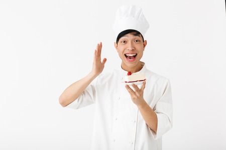 Excited asian chef wearing uniform standing isolated over white background, showing piece of cake on a plate