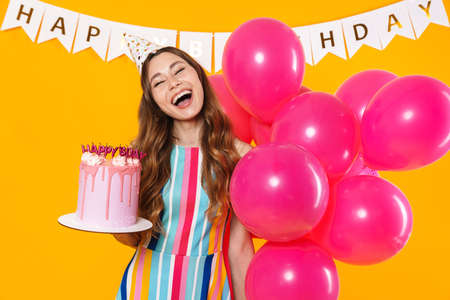 Imagem de mulher bonita excitada no cone do partido posando com balões cor-de-rosa e torte do aniversário isolado sobre o fundo amarelo - 153160785
