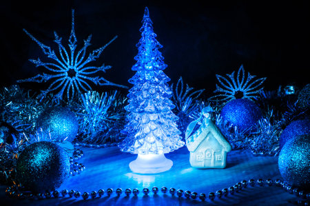 Icy christmas tree glowing with blue light on a blue background in the corner surrounded with little white house blue balls silver tinsel and snowflakes light brush