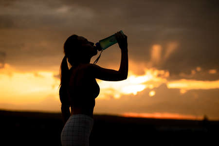 Silhouette of girl in sportswear drinks water from a bottle at sunset Banco de Imagens
