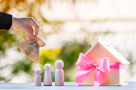 Un homme d'affaires tient à la main un sac d'argent pour sélectionner ou donner à un modèle de maison attaché avec un ruban rose et une famille de bois sur bokeh dans le parc public, l'achat d'une nouvelle maison ou d'un bien immobilier en cadeau au concept de l'être aimé.