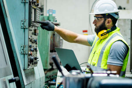 Mechanical engineer is pushing the button of the control panel while he is holding the laptop to check the list to set up in the control room at the factory