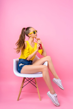 Adorable girl in sunglasses sitting in chair in studio holding bottle and drinking beverage through straw on pink background Banco de Imagens