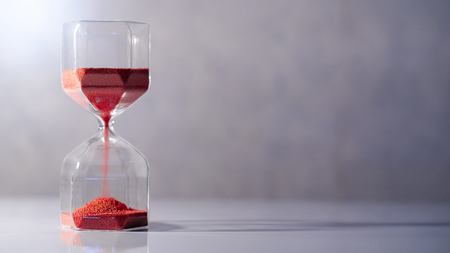 Red Sand Running Through The Shape Of Modern Hourglass On White Table.time Passing And Running Out Of Time. Urgency Countdown Timer For Business Deadline Concept With Copy Space