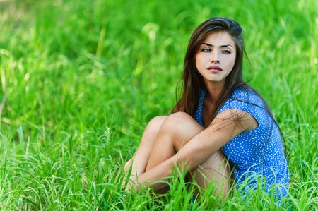 sad lonely: background summer green grass young charming, sad, long-haired in blue dress woman sitting hugging legs Stock Photo