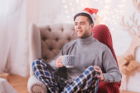 New Year time. Handsome nice happy man sitting in a chair and holding a tea cup while wearing a Santa hat
